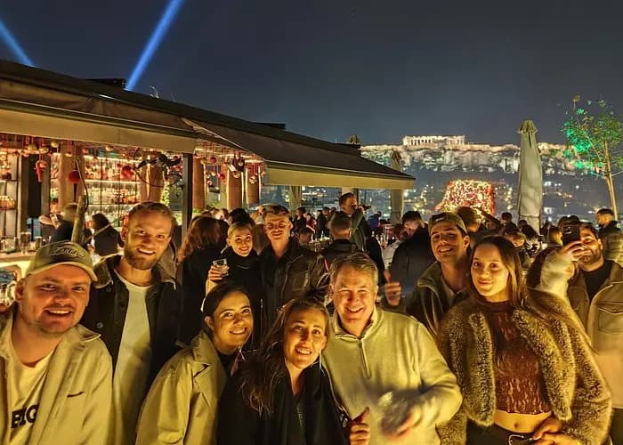 Group of international travelers enjoying rooftop bar with Acropolis views at Athens rooftop pub crawl